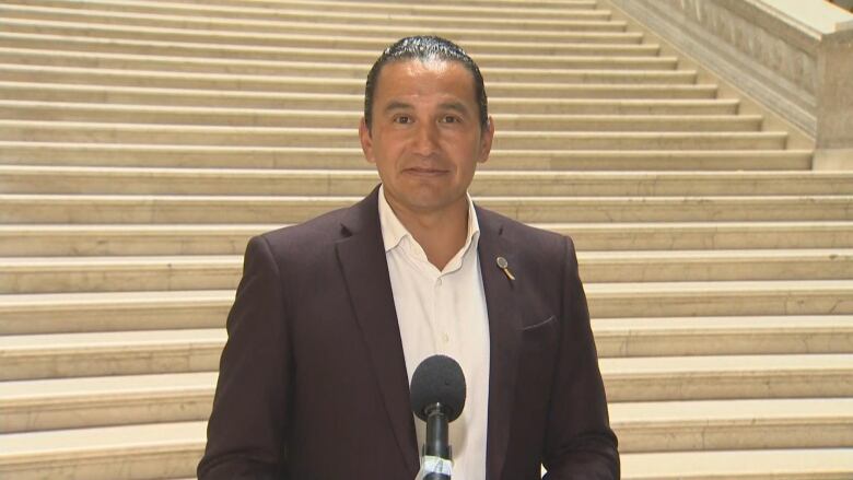 A man wearing a dark suit jacket and a white shirt stands behind a microphone in front of a white stone staircase.