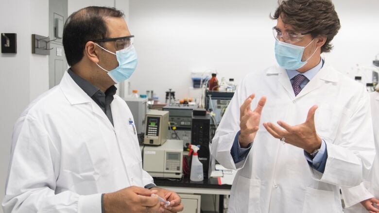 Two men wearing lab coats, face masks and safety glasses in conversation. One man holds a vaccine needle.