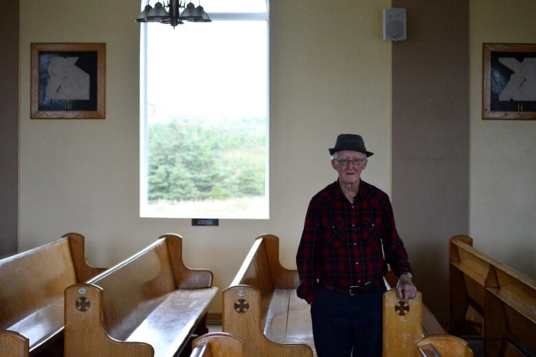 An elderly man stands in a dark church with his hands on a pew.