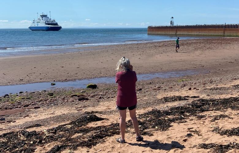 Woman stands on sandy beach taking photo of an approaching ferry. 