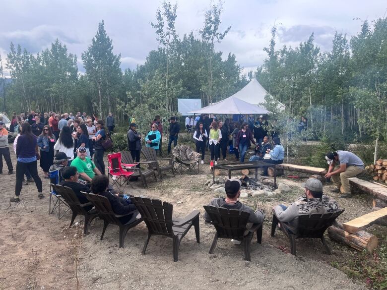 A group of people sit around a fire outdoors. It's a grey day and there are trees behind them.