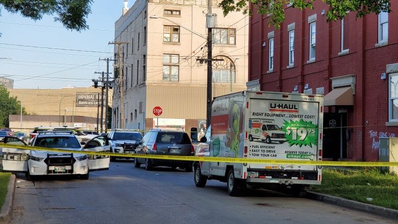 Yellow police tape stretches across a city street, with a red brick apartment building on the right. Several police cars are seen behind the tape.