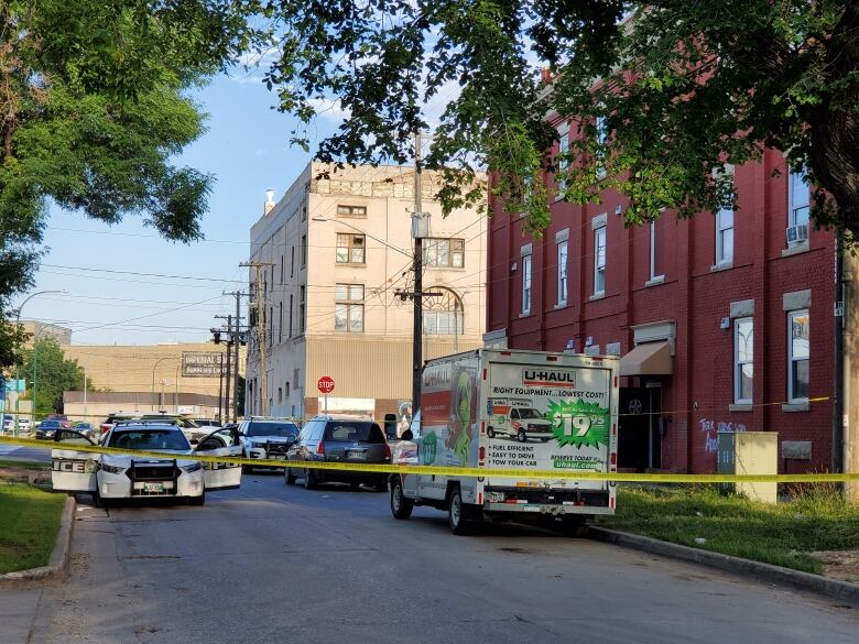 Yellow police tape stretches across a city street, with a red brick apartment building on the right. Several police cars are seen behind the tape.