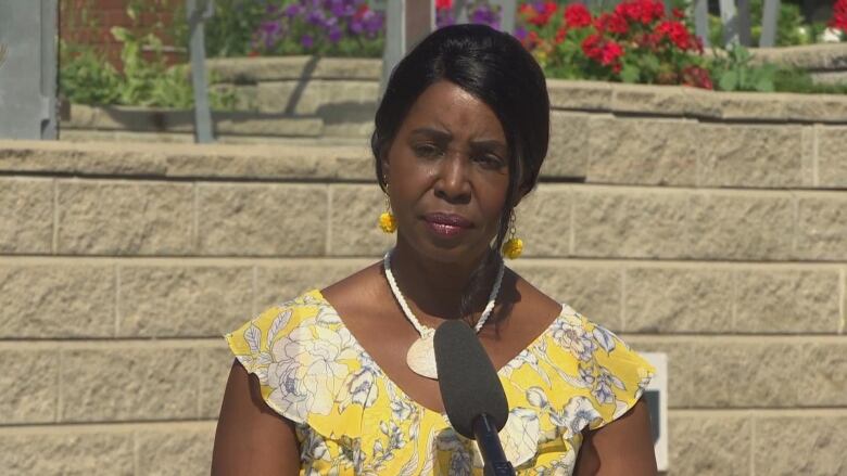 A Black woman wearing a yellow dress stands in front of a grey brick wall.