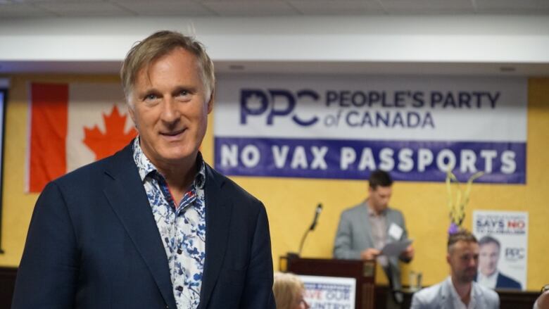 A man with grey hair wearing a white and blue shirt under a suit jacket stands in front of two signs, one that says 