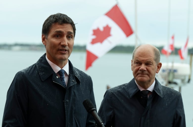 Two men wearing suits stand near a microphone in the rain. Canadian flags are in the background. 