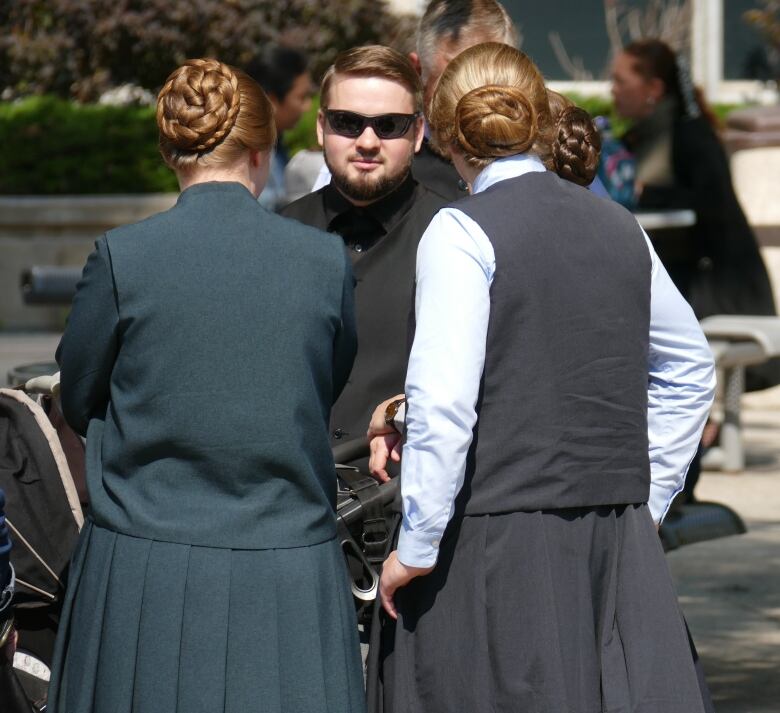 A man in sunglasses stands behind two women wearing buns in their hair and long, pleated skirts.