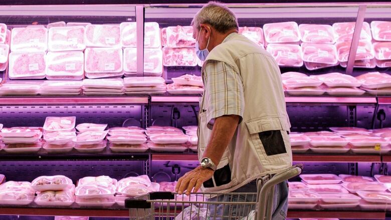 A man wearing a face mask stands behind a shopping cart, looking at packages of meat on refrigerated shelves. 