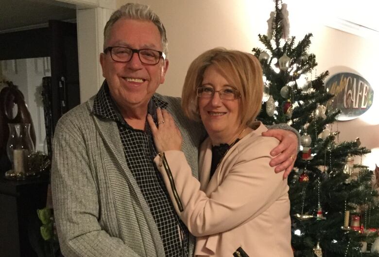 A man with grey hair and glasses stands with his arm around a woman standing beside him. They are both looking at the camera and smiling, standing in front of a Christmas tree.