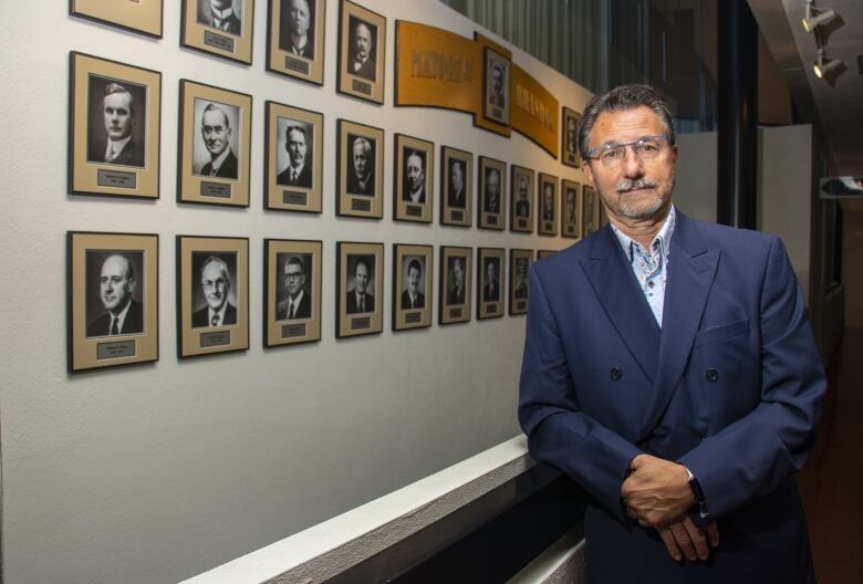 A man in a blue suit leans against a wall with multiple photo portraits on it.