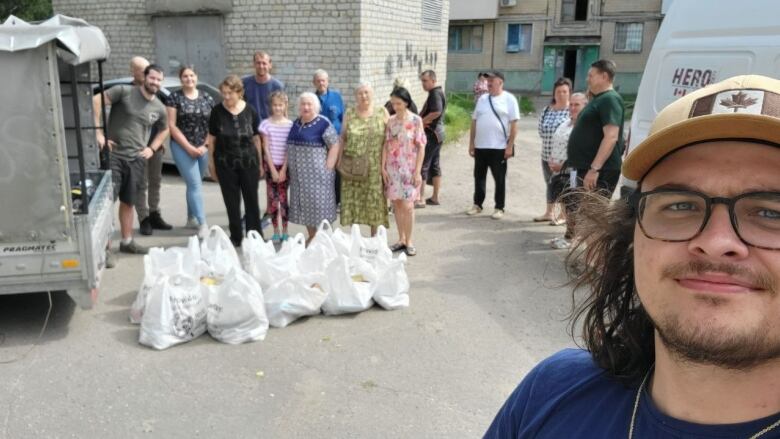 Alex stands in front of several people standing beside a large van, white bags at their feet.