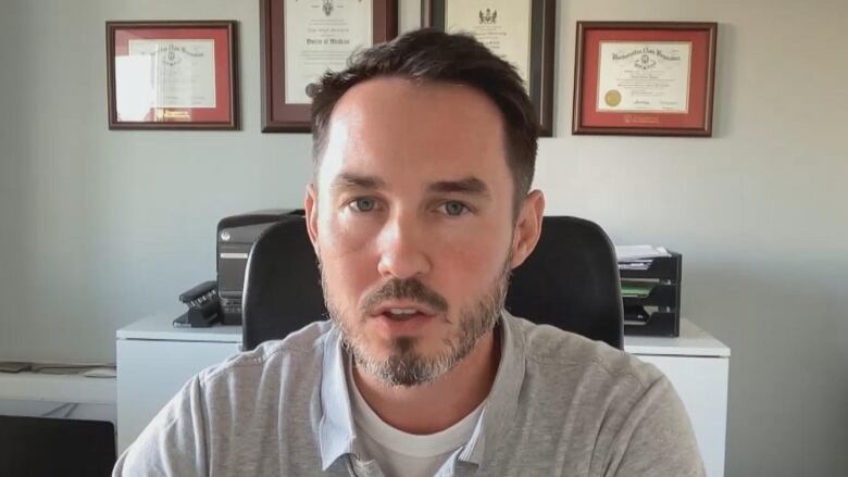 Man sits at desk with medical diplomas behind him. 