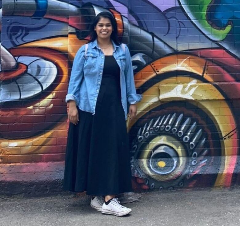 A young woman wearing a jean jacket poses in front of a graffiti decorated wall.