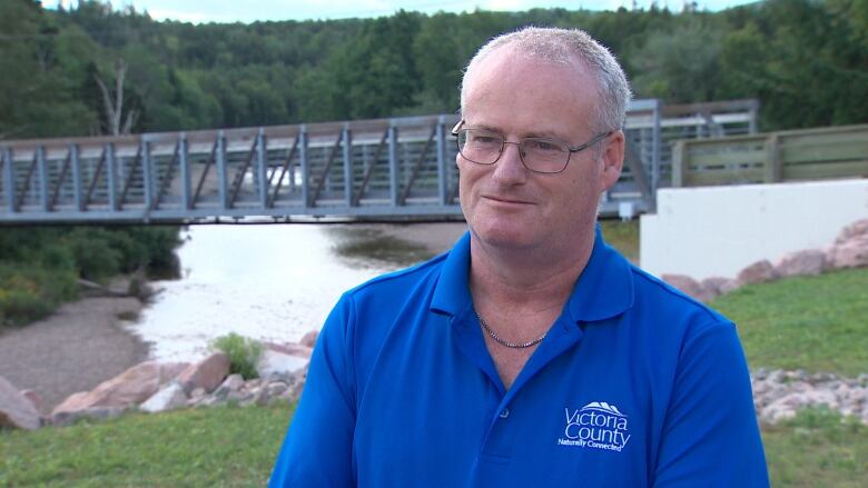 A man stands in front of a brrok with a bridge over it, with grey hair and glasses and wearing a blue shirt with a Victoria County logo on it.