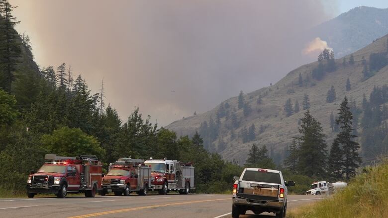 Plumes of smoke rise from a hilltop behind a highway with firetrucks and utility vehicles on it.