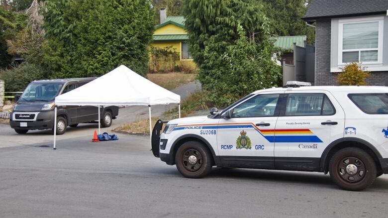 Police vehicles surround a white tent with orange cones and some clothes under it on a suburban street.