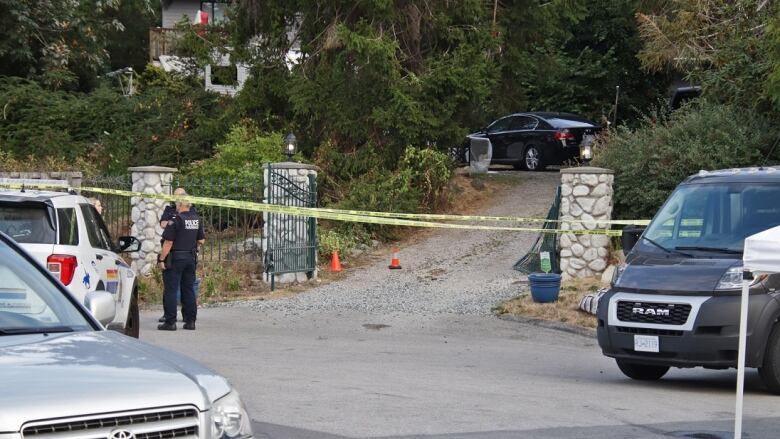 Crime tape is seen blocking off a house gate, with police officers and vehicles surrounding it.