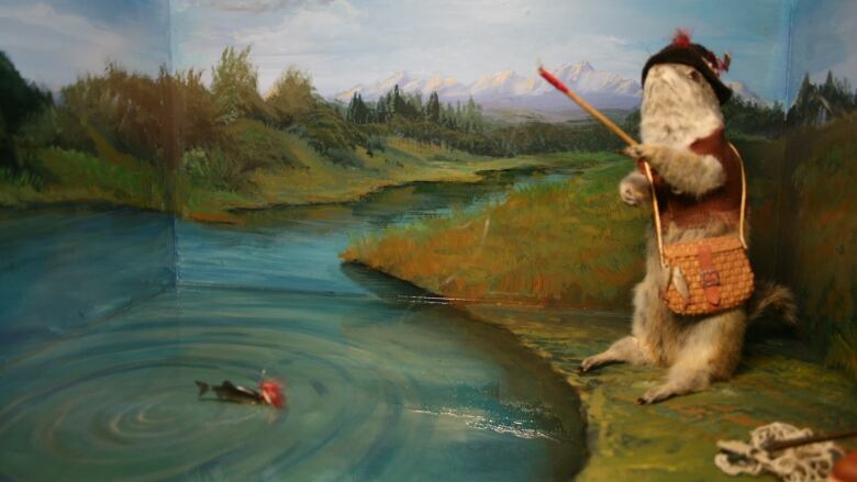 A gopher dressed as a fisherman is depicted as hooking a fish in a quiet pond in a diorama.