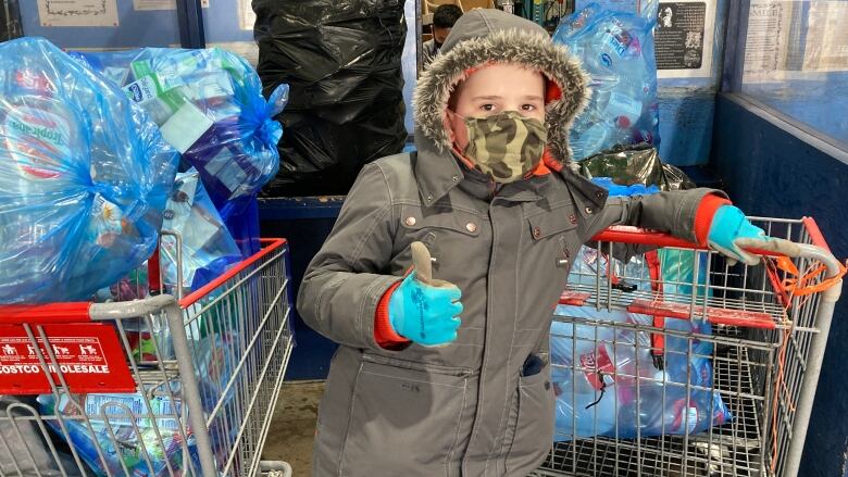 Boy wearing medical mask and warm parka gives a thumbs up while standing next to bags of refundable bottles.