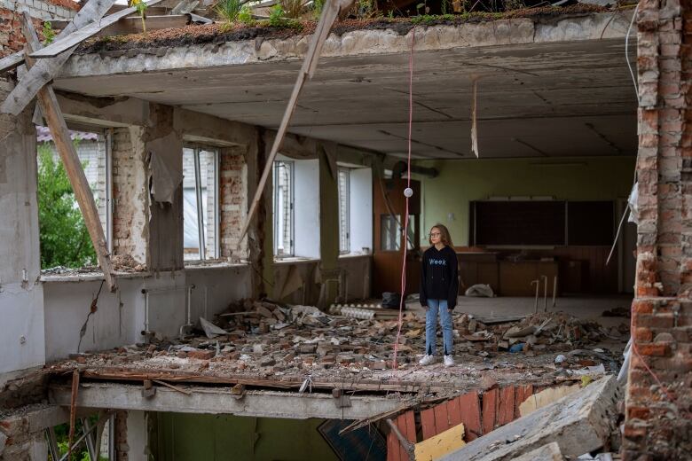 A young girl stands in the rubble of a former classroom which is missing an entire exterior wall. 