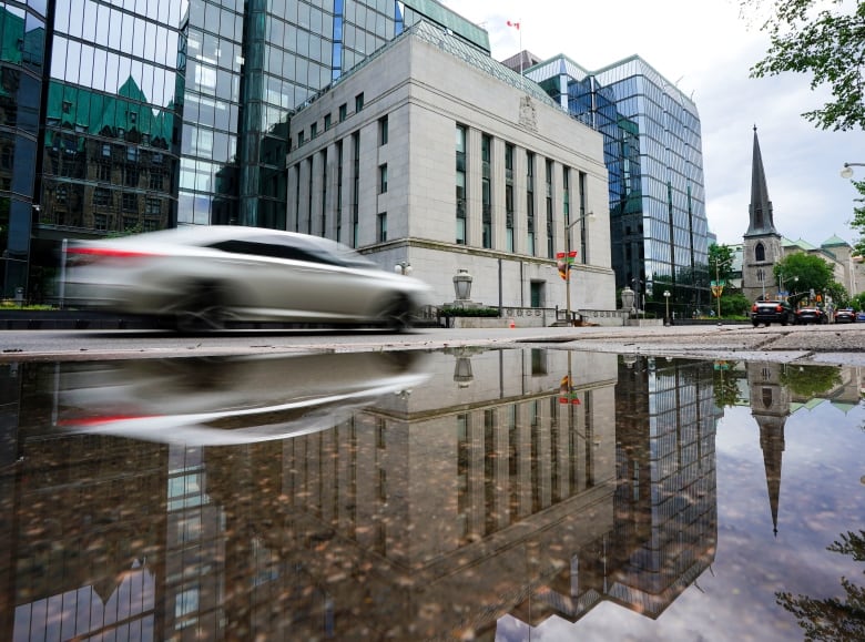 A car drives past a large stone building, flanked by larger glass office buildings. 