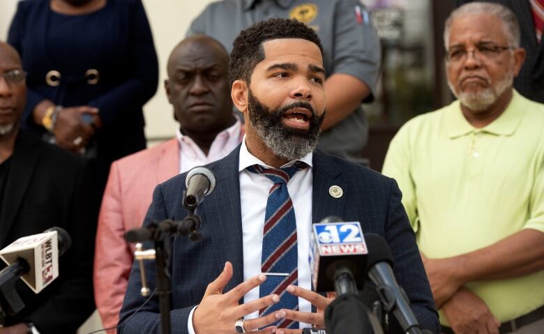 A man in a suit and tie speaks in front of several microphones from different news agencies while several people with serious expressions on their faces stand behind him. 