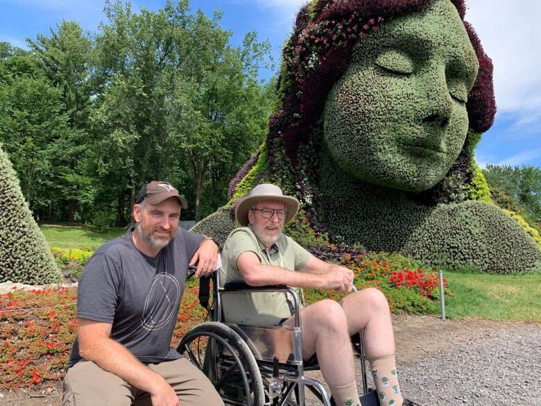 Two men, one in a wheelchair, are seen in front of a plant sculpture.