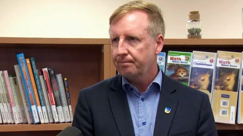 A man wearing a blue blazer and a blue unbuttoned shirt stands in front of a bookcase in a school classroom. 