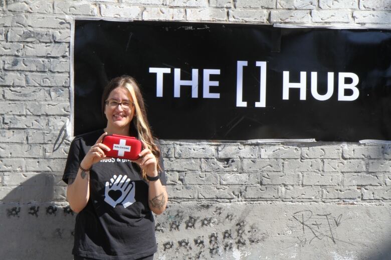 Olivia Mancini stands in front of a sign for The Hub on a brick wall, holding up a naloxone treatment kit above their shirt which bears a logo of two hands over a heart.