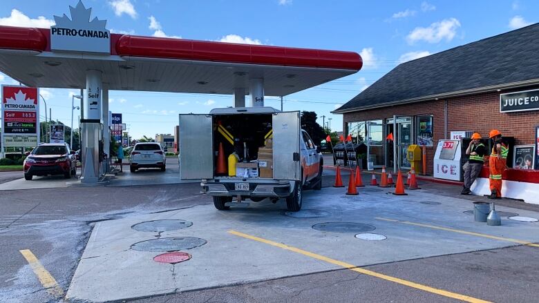 Orange hazard cones stand near the covers of in-ground gasoline holding tanks at a gas station as cars fill up nearby. 