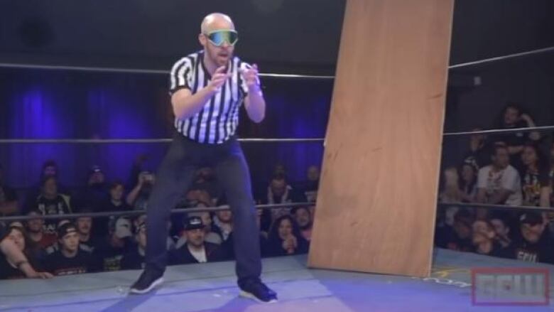 A wrestling referee is standing with thick tinted glasses watching a match between two invisible opponents in the ring.