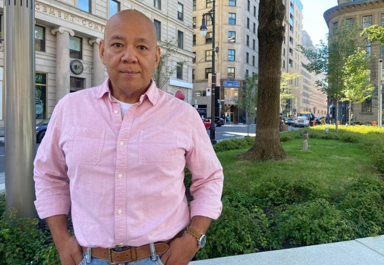 bald Asian man wearing a pink button-up shirt and blue jeans with a brown belt and watch standing outside on a summer day