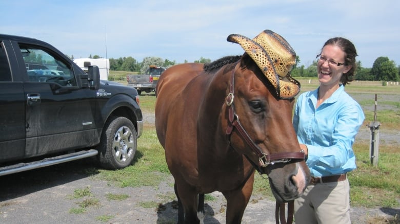 A woman laughs as she stands beside a horse with a cowboy hat perched on its head.