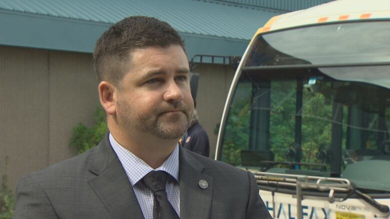 A white man in a grey suit and tie stands in front of a transit bus