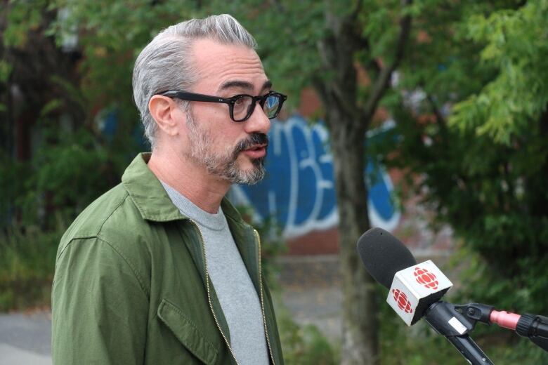 Man with grey hair and beard and black-rimmed glasses speaks into CBC mic.