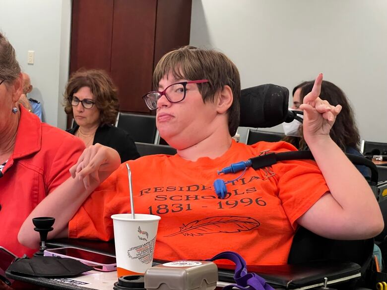 A woman in an orange t-shirt wearing glasses, sitting in a wheelchair.