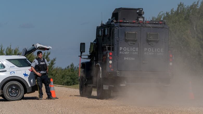 A large black armoured vehicle drives on a dirt road past an RCMP SUV with an officer standing outside.