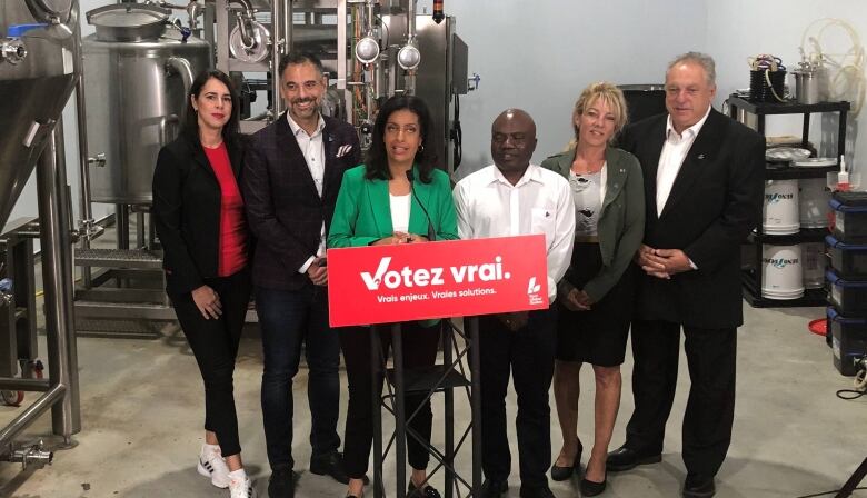 Six people stand near a podium in front of brewing vats.