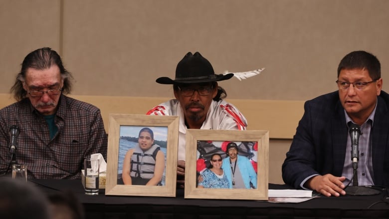 Three men sit at a conference table, on which sit framed photos of three other people. 