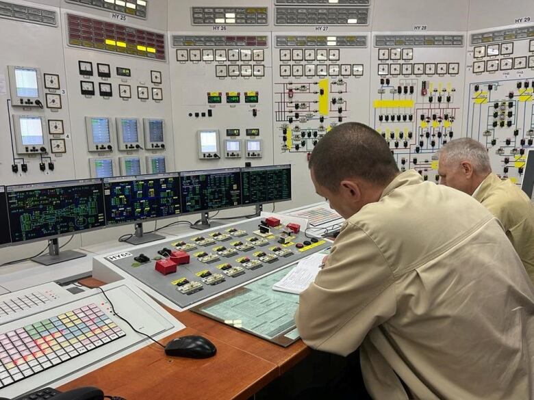 Two men in beige shirts sit at a table and wall covered with indicators and controls.