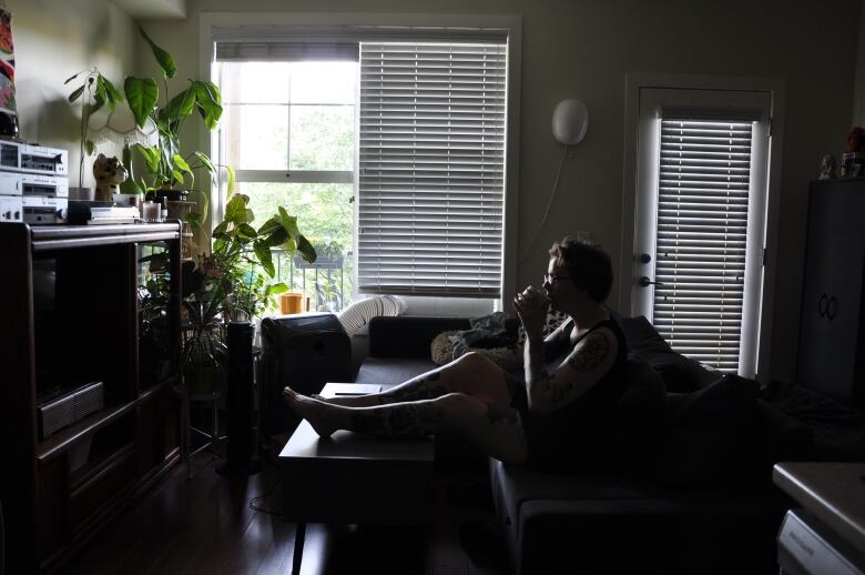 A woman sits on a couch in front of a bright window sipping a drink. 