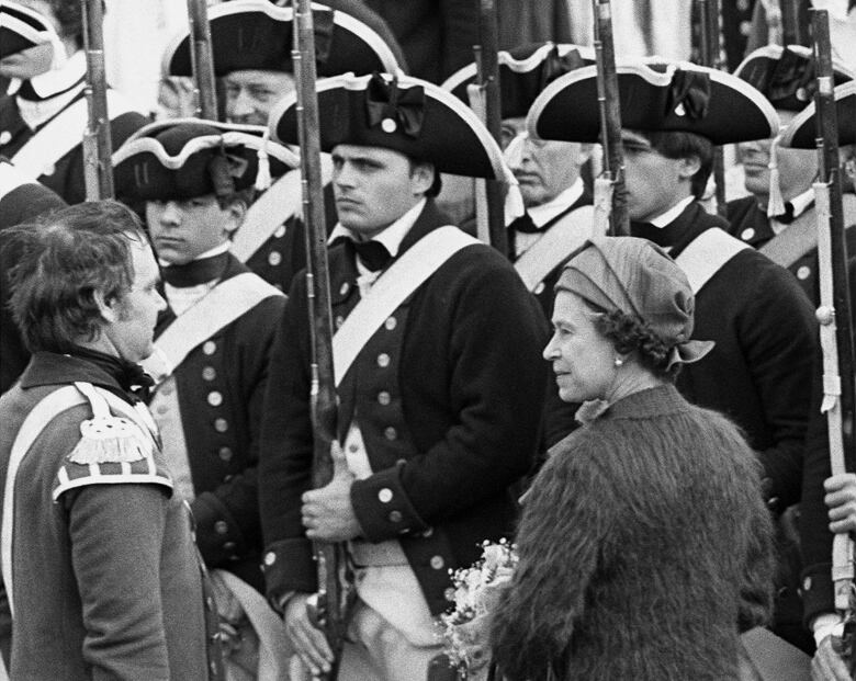 The Queen watches as soldiers in old-fashioned gear march by her.