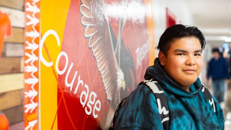 A young man wearing a blue hoodie and a backpack stands in front of a colourful mural.