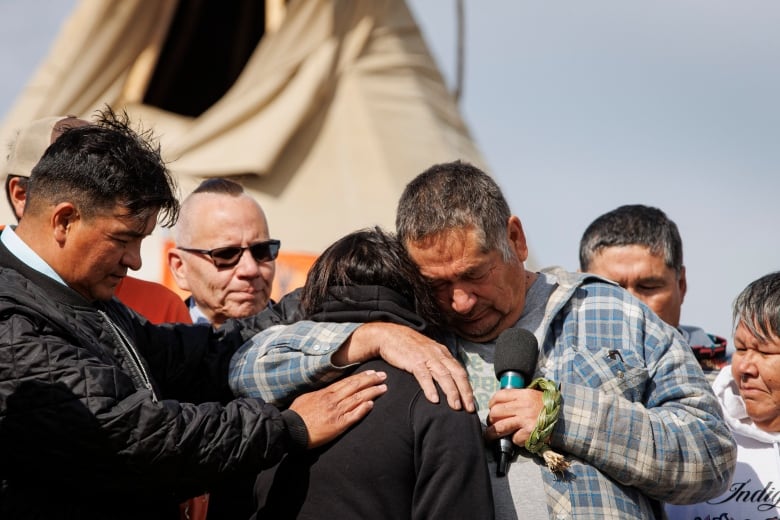 A man puts his arm around a woman who has her head down, while others stand by. Behind them stands a teepee.