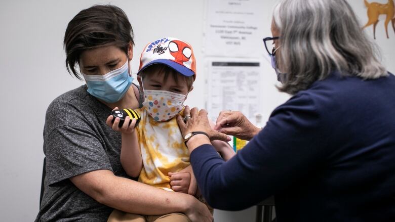 A child gets a needle while holding a bumblebee toy and sitting with an adult.