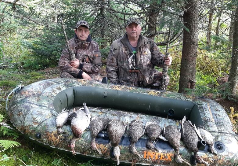 Two men dressed in camouflage hunting gear stand in the woods behind a camouflage-patterned inflatable raft, on which nine dead ducks are spread.