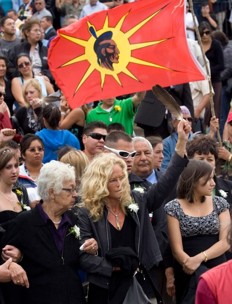 a woman holds a feather near a Mi'kmaq flag at a large gathering of people outside a church. 