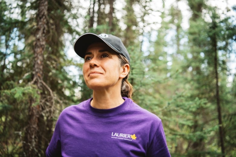 A woman in a purple shirt and ball cap looks up beyond the camera in the midst of a forest.