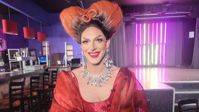 A drag queen with dramatic orange hair and sparkling necklace smiles at the camera.