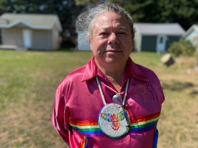 A person wearing a bright red and rainbow top and a beaded necklace smiles and stands in a park.